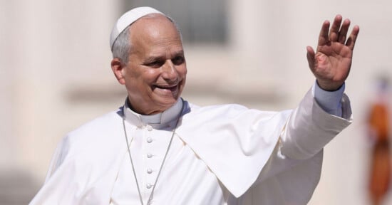 A man dressed in white papal vestments and a white zucchetto smiles and waves his left hand while looking toward the right.
