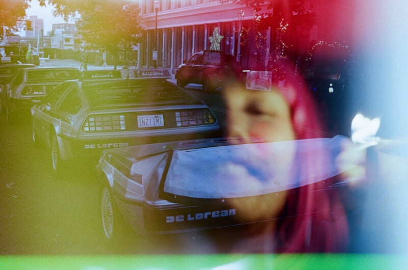 Two silver DeLorean cars are parked on a city street, partially obscured by a double-exposure overlay of a person's face.