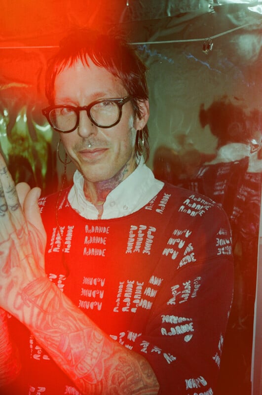 A person with a neck tattoo and glasses wears a red patterned sweater over a white collared shirt, with their tattooed arm raised in the foreground.