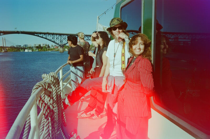 A group of people stand on the deck of a boat on a sunny day, with a large bridge visible in the background.