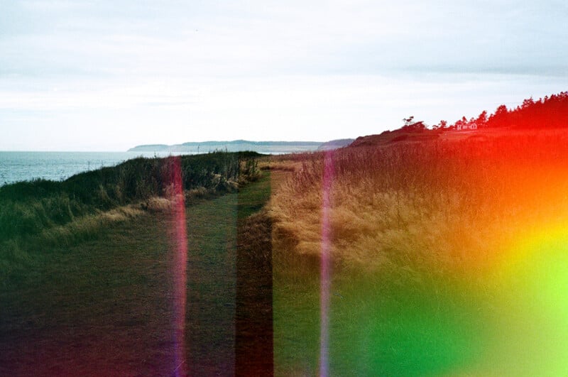 A grassy path runs along a coastal cliffside under a cloudy sky, with prominent light leaks and color distortions across the frame.