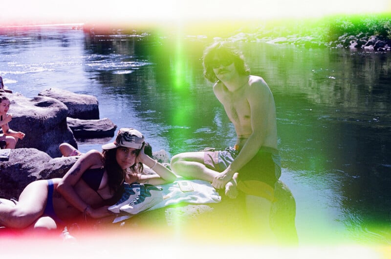 A woman in a bikini and baseball cap reclines on a rock next to a shirtless man wearing sunglasses by a river, with light leaks visible across the frame.