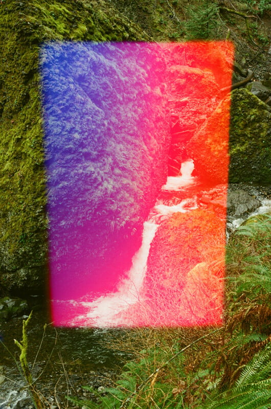 A small waterfall flows between mossy rock faces, partially obscured by a rectangular, vertical gradient of blue, pink, and orange light.