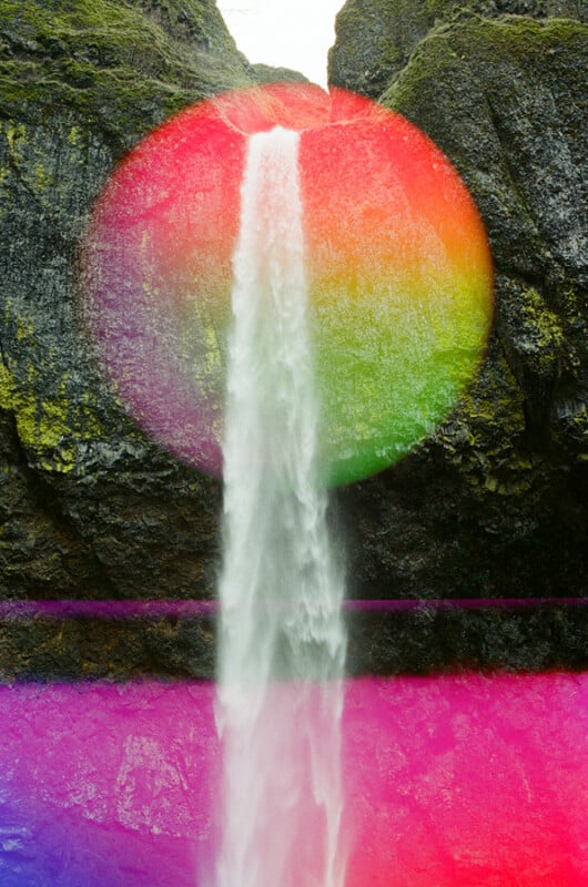 A waterfall flows down a mossy rock cliff, overlaid with a large, circular gradient of red, orange, and green, and a horizontal band of bright pink light.