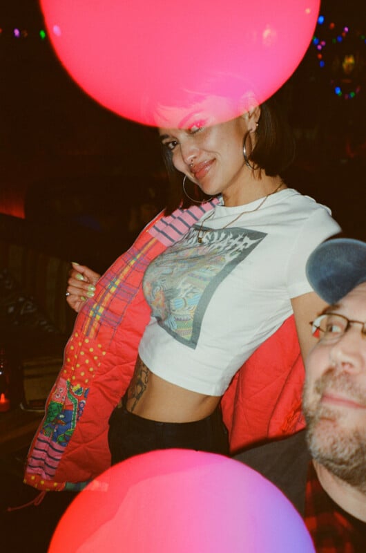 A woman with a nose ring and hoop earrings poses in a white graphic t-shirt and a colorful patterned jacket, partially obscured by two large, glowing pink circular light effects.
