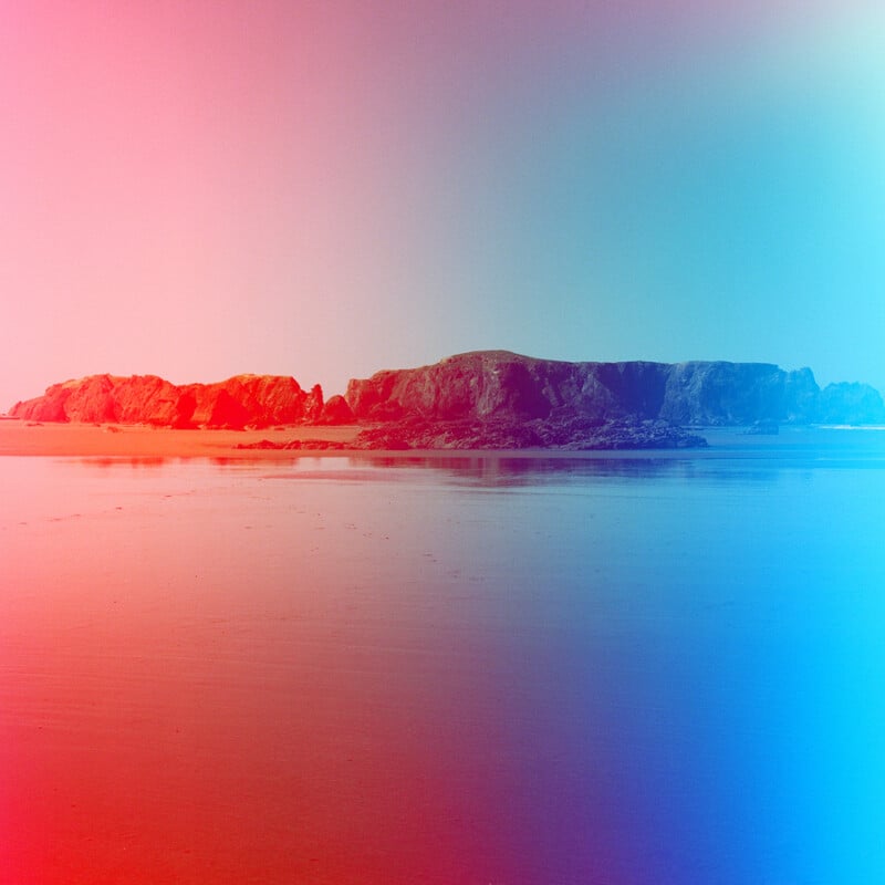 A rocky island sits in the middle of a calm, reflective beach under a sky transitioning from pink on the left to blue on the right.
