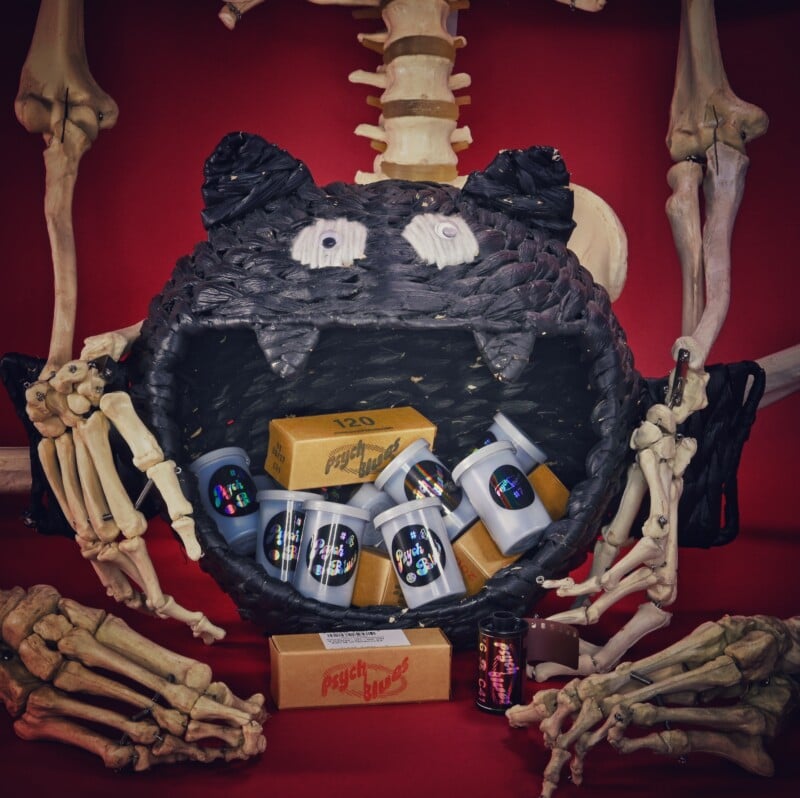 A skeleton holds a large, black, woven cat-shaped basket filled with several film canisters and boxes labeled "Psych Blues" against a red background.