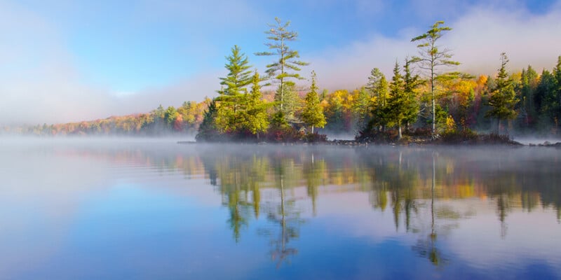 A calm lake reflects a small, tree-covered island surrounded by morning mist and autumn-colored foliage.