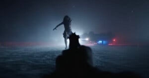 A first-person view looking down the barrel of a handgun at a woman standing in a snowy, dark landscape near an overturned car.