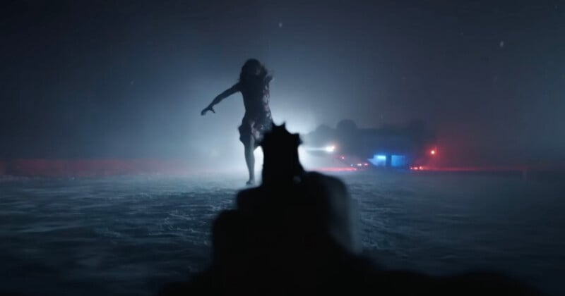 A first-person view looking down the barrel of a handgun at a woman standing in a snowy, dark landscape near an overturned car.