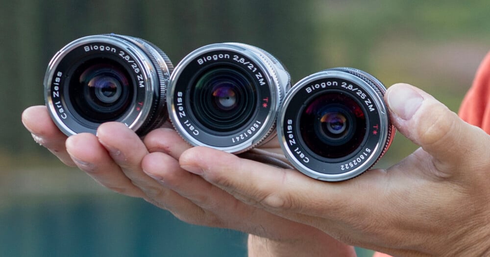 A person's hand holds three Carl Zeiss Biogon ZM lenses, labeled 2.8/28, 2.8/21, and 2.8/25, against a blurred outdoor background.