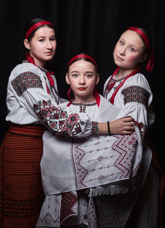 Three young girls in traditional Ukrainian clothing with red headbands pose together against a black background.