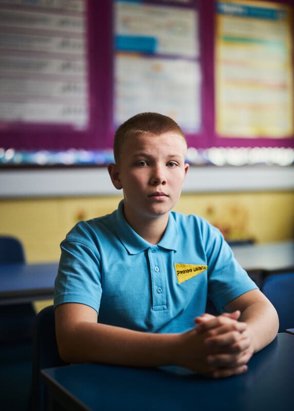 A young boy with short hair sits at a desk in a classroom, wearing a light blue polo shirt with a yellow patch on his chest.