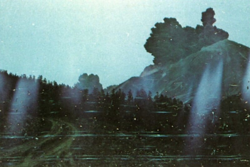 A dark, plume-like cloud rises above a forested hillside, with light streaks visible across the foreground.