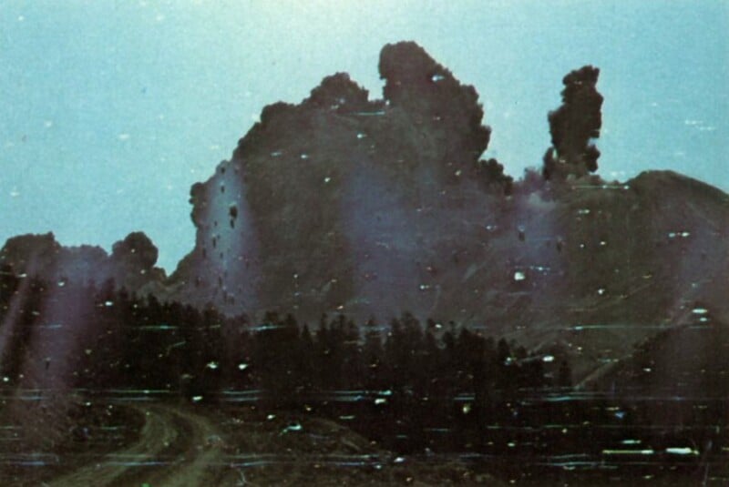 A dark, billowing cloud of smoke rises above a treeline and a dirt road, with visible horizontal scan lines across the scene.