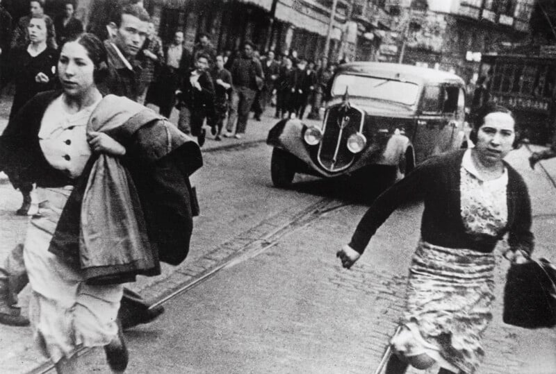 Two women run across a cobblestone street toward the camera, while a vintage car and other pedestrians move in the background.