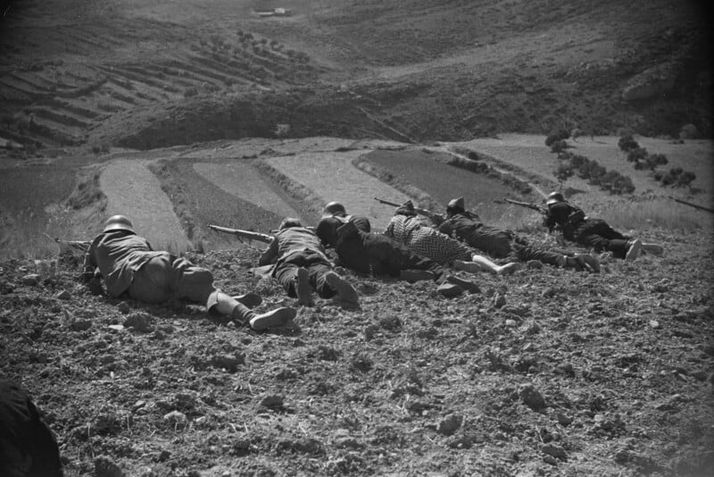 Five soldiers in helmets lie prone in a line on a dirt hillside, aiming their rifles toward the valley below.