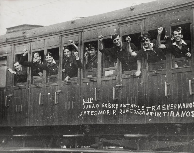 A group of men in dark uniforms and caps lean out of the windows of a wooden train carriage, raising their fists in solidarity.