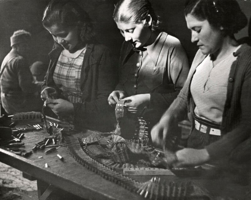 Three women work at a long table, assembling ammunition belts by hand in a dimly lit room.