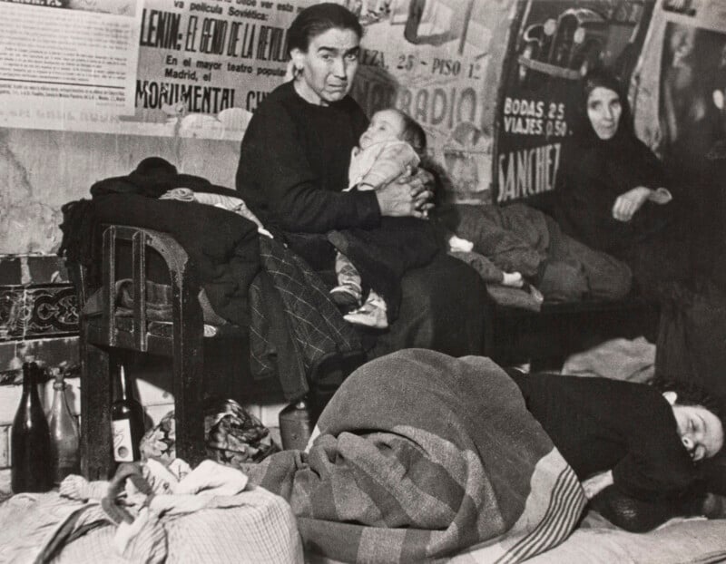 A woman holds a baby while sitting in a room with posters on the wall, as another person sleeps on the floor nearby.