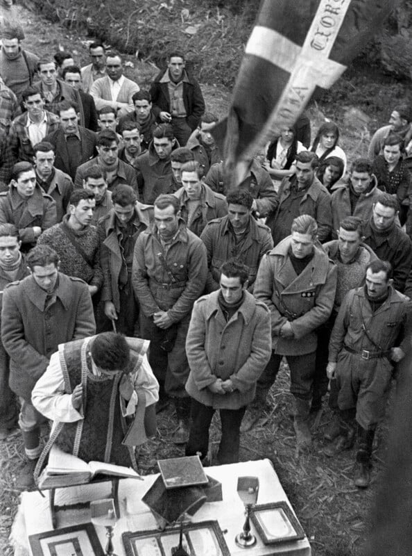 A priest conducts an outdoor religious service for a large group of men in military-style clothing, with a flag visible in the background.