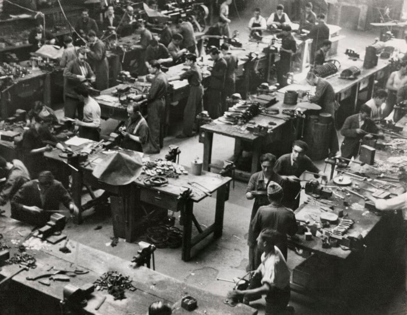 A high-angle view of a crowded industrial workshop filled with men working at individual wooden benches equipped with vises and hand tools.