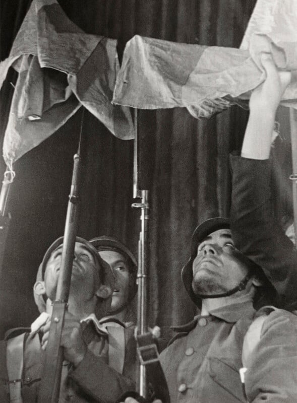 Three soldiers in helmets look upward while holding rifles and a fabric banner.