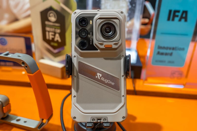 A Rugone rugged smartphone is mounted on a display stand, featuring a prominent camera module with multiple lenses and a textured light-gray back panel.