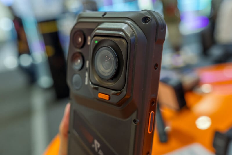 A close-up, angled view of the back of a rugged smartphone featuring a large, prominent circular camera lens, two smaller lenses, and an orange accent button.