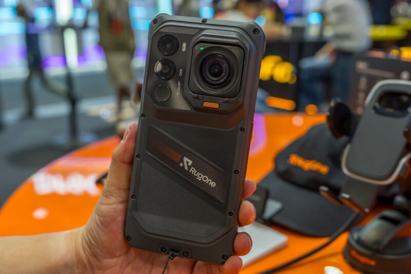 A hand holds a rugged, dark gray smartphone with a prominent camera module and the brand name "RugOne" printed on the back.