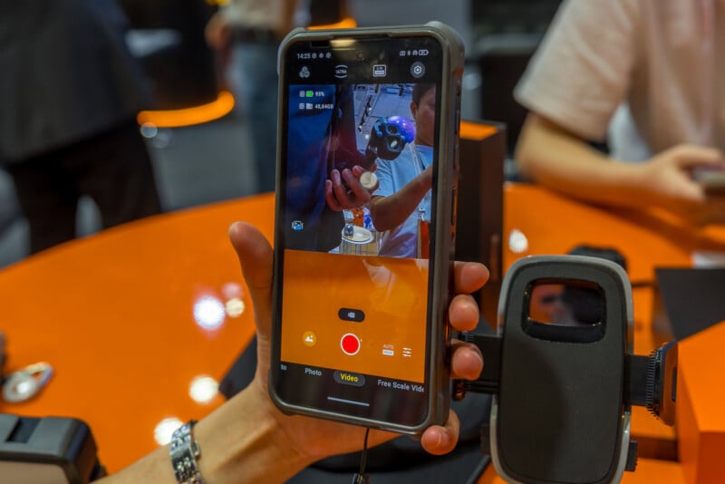 A hand holds a rugged smartphone displaying its camera interface, with a car phone mount positioned to the right on an orange table.