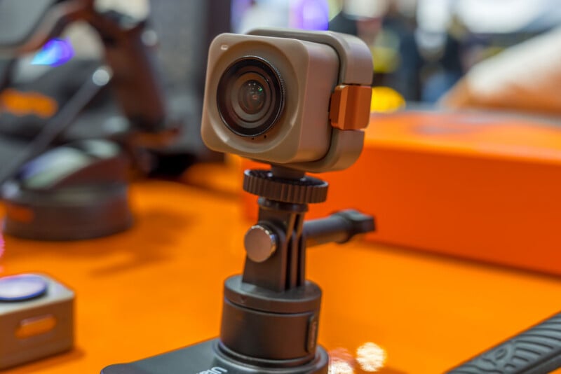 A small, square, tan-colored action camera mounted on a black tripod stand sits on an orange surface.
