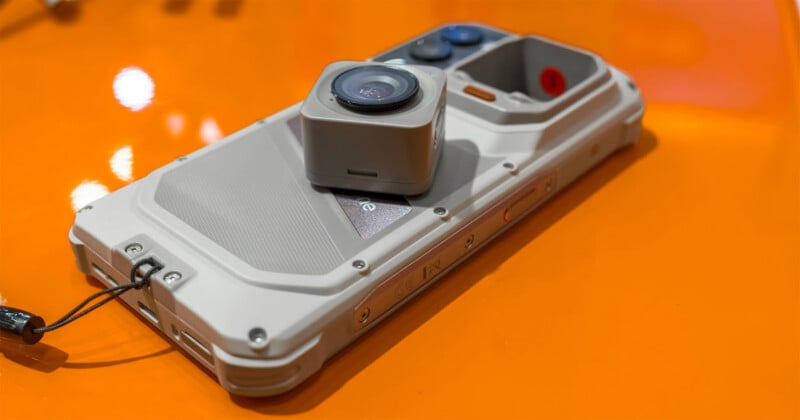 A light-colored, rugged smartphone with a detachable square camera module resting on its back, set against a bright orange surface.