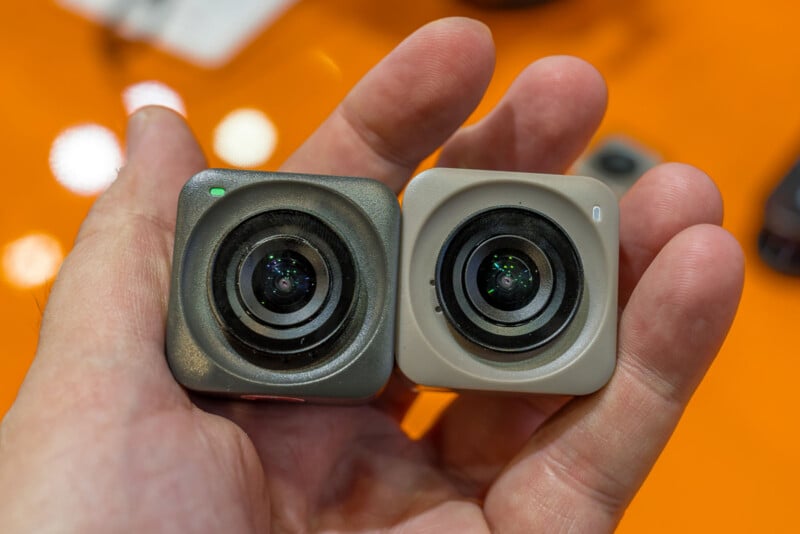 A hand holds two small, square action cameras side-by-side against an orange background.