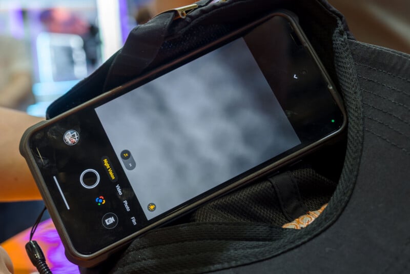 A smartphone in a protective case rests inside a black baseball cap, displaying a camera interface with a blurred, gray background.