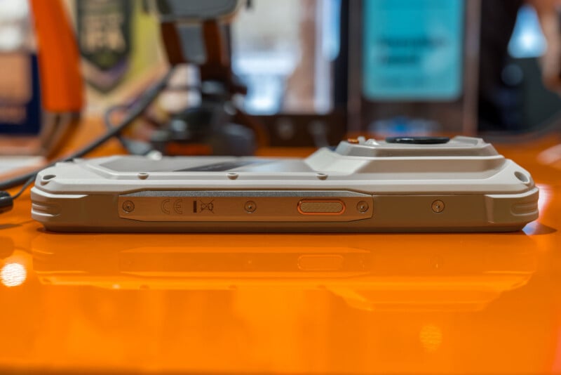 A side view of a rugged, light-colored smartphone resting on a reflective orange surface, showing a metallic side panel with screws, regulatory markings, and a textured power button.