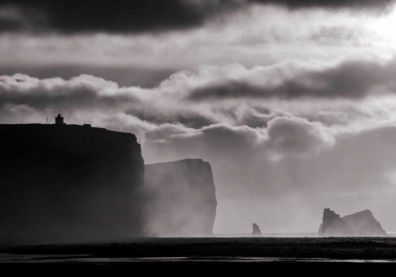A black-and-white landscape featuring dark, silhouetted cliffs rising above a beach, with a lighthouse visible on the summit.