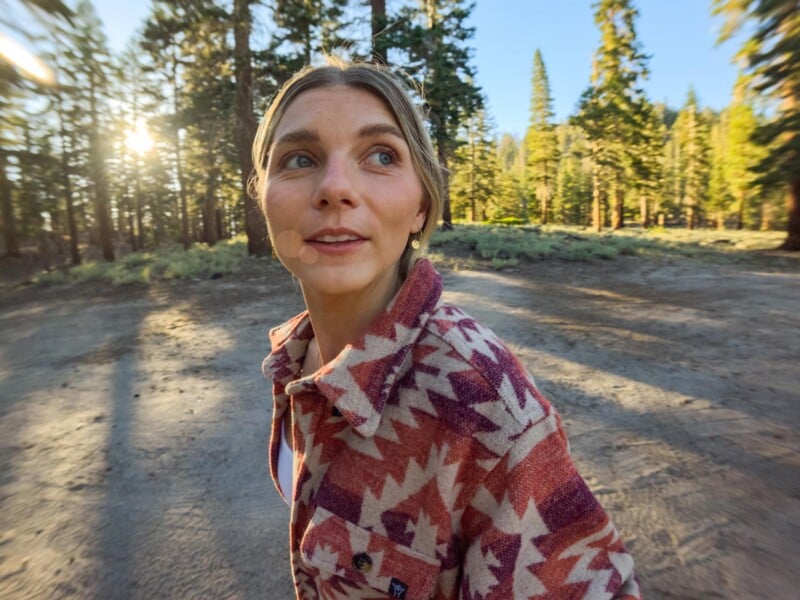 A woman with blonde hair looks to the side while wearing a patterned, button-down shirt in a sunlit forest.