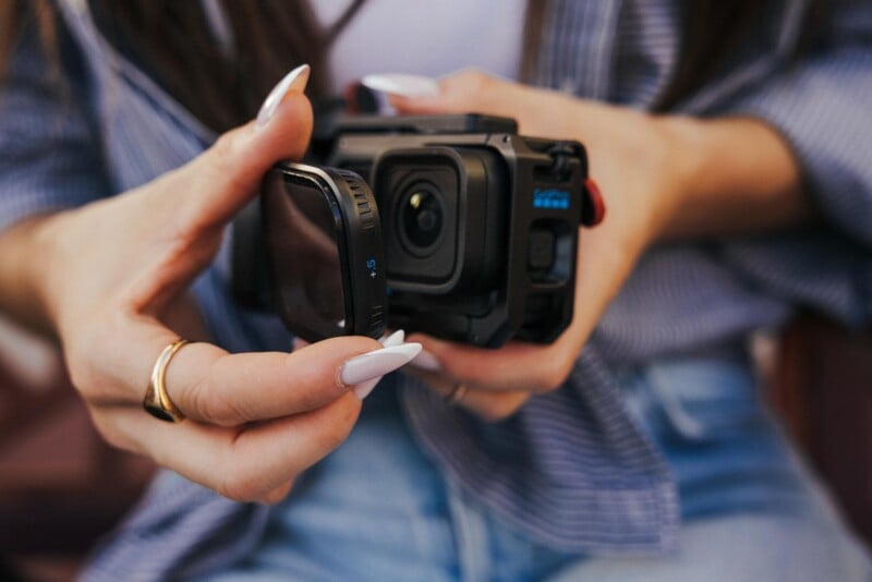 A person with long, white-tipped fingernails holds a square camera lens filter labeled "+.5" in front of a black action camera.