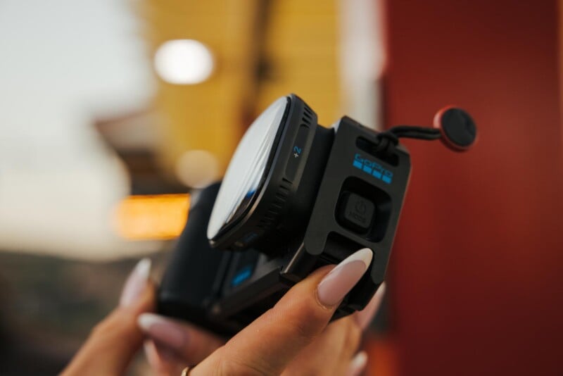 A person holds a GoPro camera with a macro lens attachment, showing the power button and a small red tether.