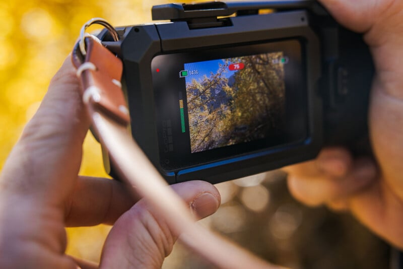 A person holds a black action camera in a protective housing, displaying a live view of yellow autumn trees on its rear screen.