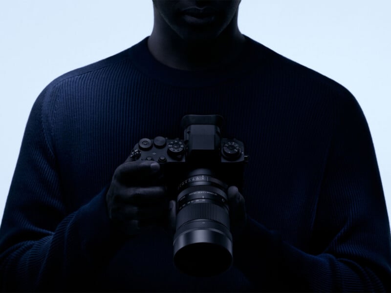 A person wearing a dark ribbed sweater holds a camera with a zoom lens against a bright, backlit background.