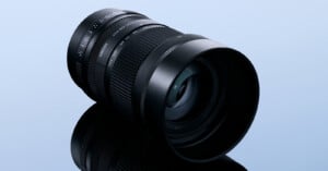 A black camera lens with a zoom ring and aperture markings sits on a reflective surface against a light blue background.
