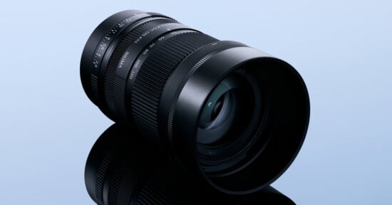 A black camera lens with a zoom ring and aperture markings sits on a reflective surface against a light blue background.