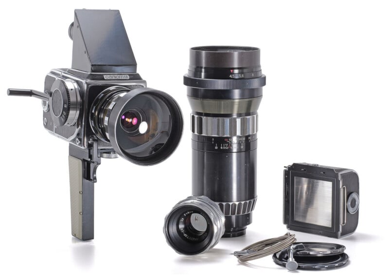 A Salyut-S medium format camera with a waist-level viewfinder, a large telephoto lens, a smaller Industar-29 lens, a film back, and a cable release on a white background.