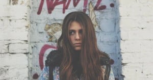 A young woman with long brown hair looks to the side while standing in front of a white brick wall with graffiti.