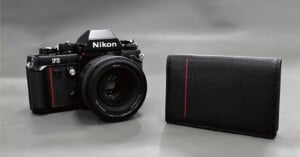 A black Nikon F3 film camera with a lens sits next to a black leather wallet featuring a vertical red stripe on a gray background.