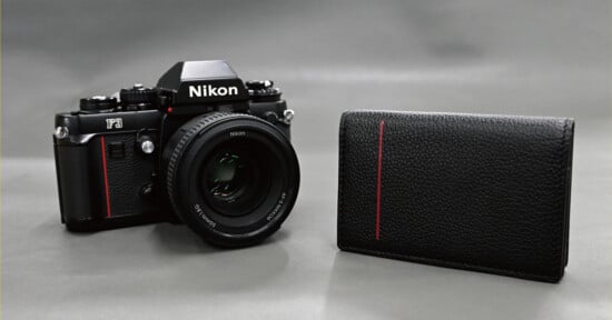 A black Nikon F3 film camera with a lens sits next to a black leather wallet featuring a vertical red stripe on a gray background.
