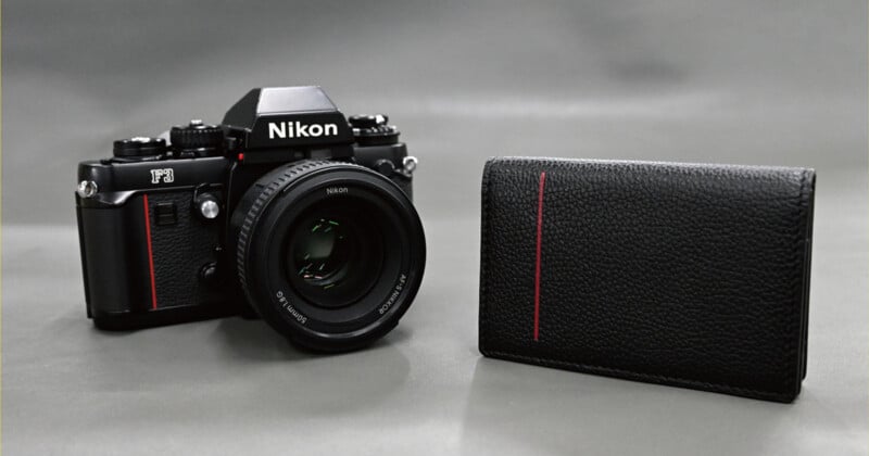 A black Nikon F3 film camera with a lens sits next to a black leather wallet featuring a vertical red stripe on a gray background.