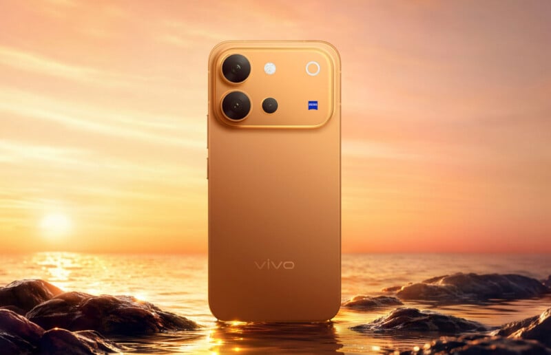 A gold-colored smartphone stands upright in shallow water against a sunset background, featuring a prominent camera module with a ZEISS logo and the word "vivo" printed on the back.